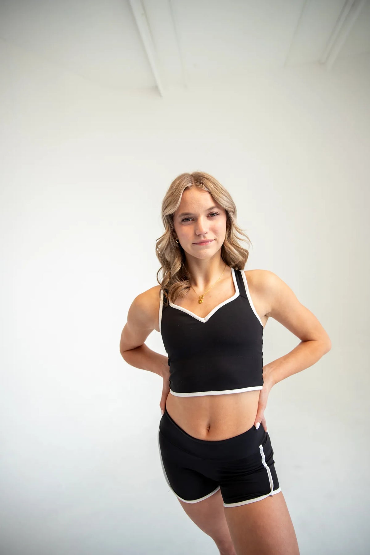 A woman modeling a black athletic crop top and matching black shorts with white trim, standing against a plain white studio wall.