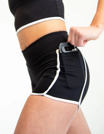 Black athletic shorts with white contrast piping and a side pocket, shown on a model with a smartphone inserted into the pocket.