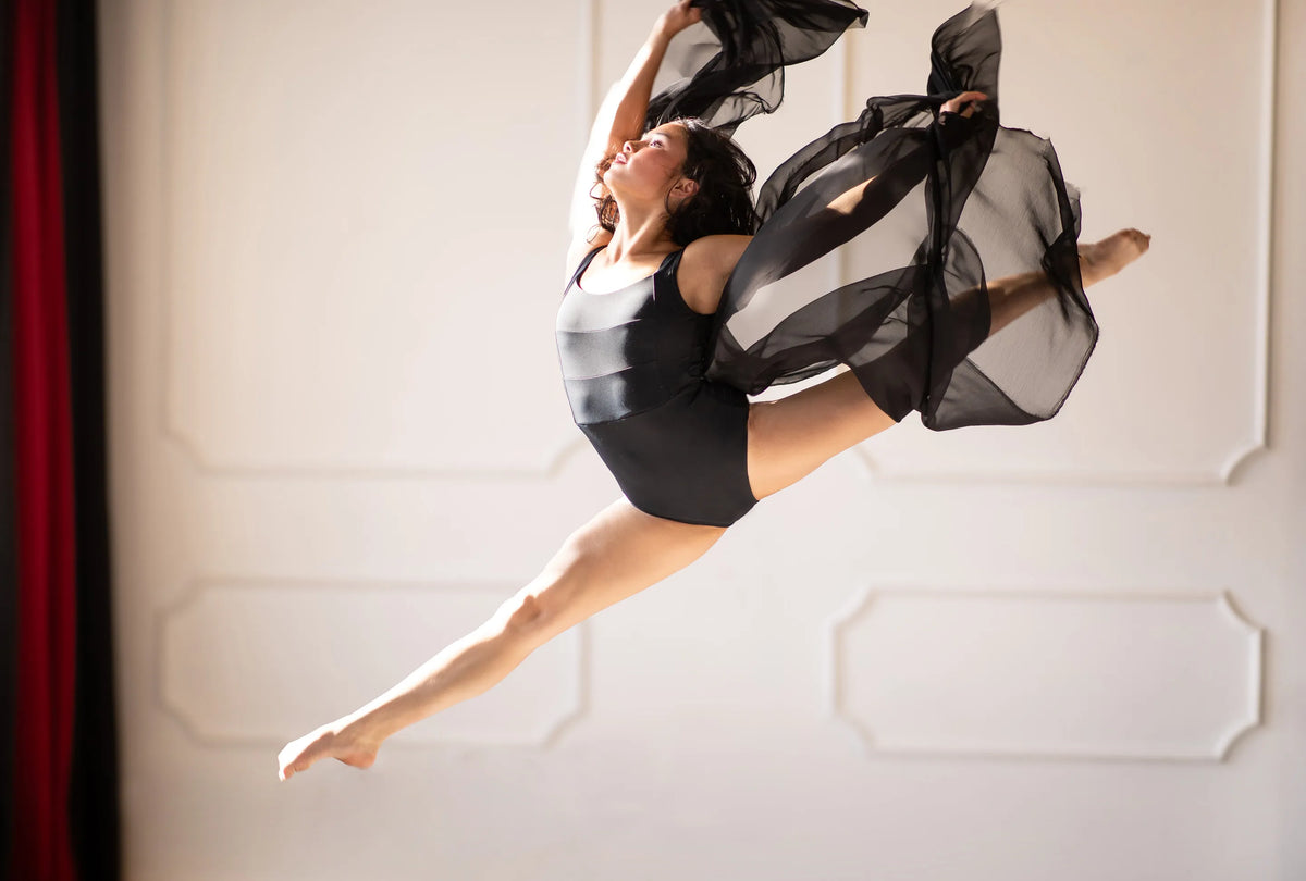 A dancer wearing a black Signature Leo leotard leaps mid-air with a sheer black scarf trailing behind her.